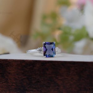 Puede incluir: Un anillo de plata con una gran gema rectangular azul-púrpura como pieza central. Dos gemas claras más pequeñas flanquean la piedra principal. El anillo está sobre una superficie marrón oscuro, con un fondo floral borroso.