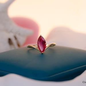 Peut inclure: Une bague en or ornée d'une pierre précieuse rouge foncé de forme marquise. La bague est posée sur une surface bleu canard foncé, avec un arrière-plan flou et doux. Le corps de la bague épouse la pierre.