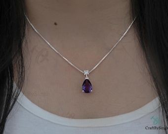 May include: A silver necklace with a pear-shaped, deep purple gemstone pendant. The pendant is suspended from a small, square-cut clear stone. The chain is a delicate, square-link style.
