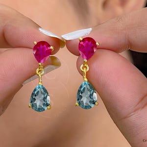 Op de afbeelding: Een paar oorhangers met een traanvormige blauwe edelsteen en een kleinere roze edelsteen erboven. De oorhangers zijn van goud.