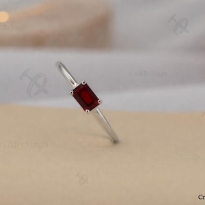 May include: A silver ring with a rectangular, deep red gemstone. The slender band holds the gemstone horizontally. The ring is shown against a neutral backdrop, emphasising its simple elegance and design.