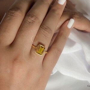 May include: A gold ring with a square-shaped, yellow gemstone. The ring is on a person's finger.