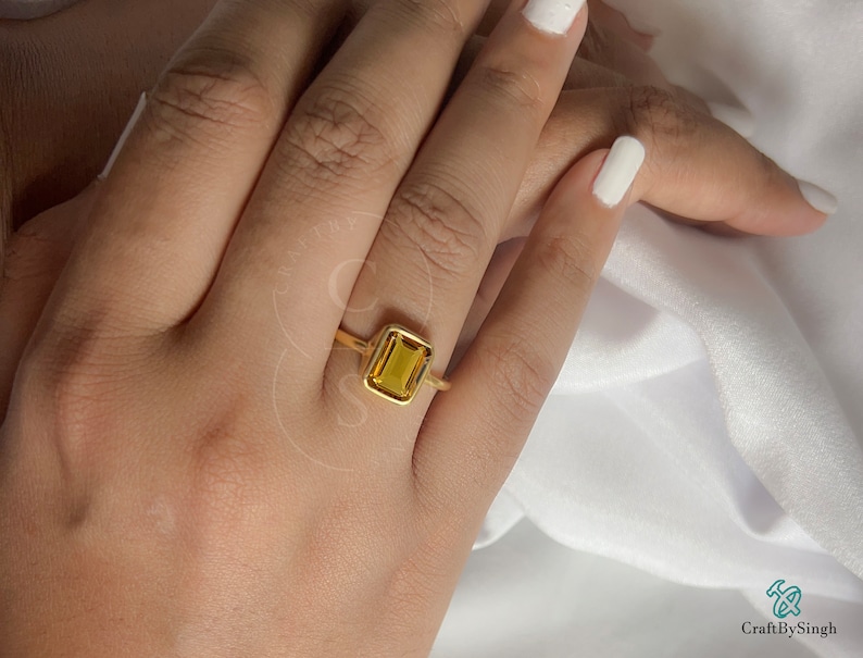 May include: A gold ring with a square-shaped, yellow gemstone. The ring is on a person's finger.
