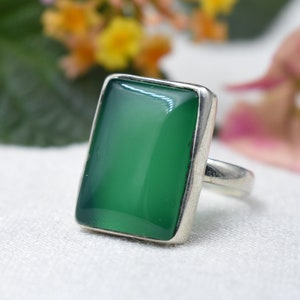 Puede incluir: Un anillo de plata con una piedra verde pulida, rectangular y grande. La piedra está engastada en una sencilla banda de plata. El anillo se muestra sobre un fondo blanco suave con acentos florales borrosos.
