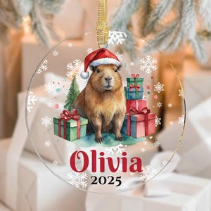 May include: A clear, round Christmas ornament featuring a capybara wearing a Santa hat, surrounded by wrapped gifts and snowflakes. The name "Olivia" and the year "2025" are printed in red. A gold ribbon is attached for hanging.