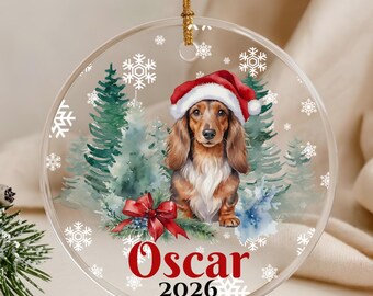 May include: A round clear acrylic ornament featuring a long-haired dachshund wearing a red and white Santa hat. The design includes watercolor evergreen trees, a red bow, white snowflakes, and the red text Oscar 2026 with a gold string.