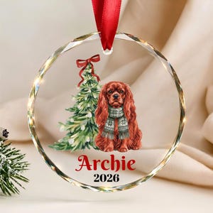 May include: A circular clear glass ornament with a red ribbon hanger featuring a watercolor illustration of a brown spaniel wearing a patterned scarf next to a decorated Christmas tree, with the name Archie and the year 2026 printed below.
