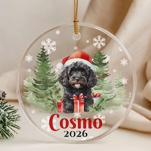 May include: A round clear acrylic ornament featuring a watercolor illustration of a black dog wearing a red Santa hat, surrounded by green pine trees and white snowflakes. Red gift boxes are at the base, with the name Cosmo and year 2026.