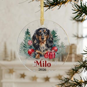 May include: A circular clear acrylic Christmas ornament featuring a long-haired dachshund illustration surrounded by green pine trees, red ornaments, and a gift box. The name Milo and the year 2026 are printed in red below the dog, with a gold ribbon.