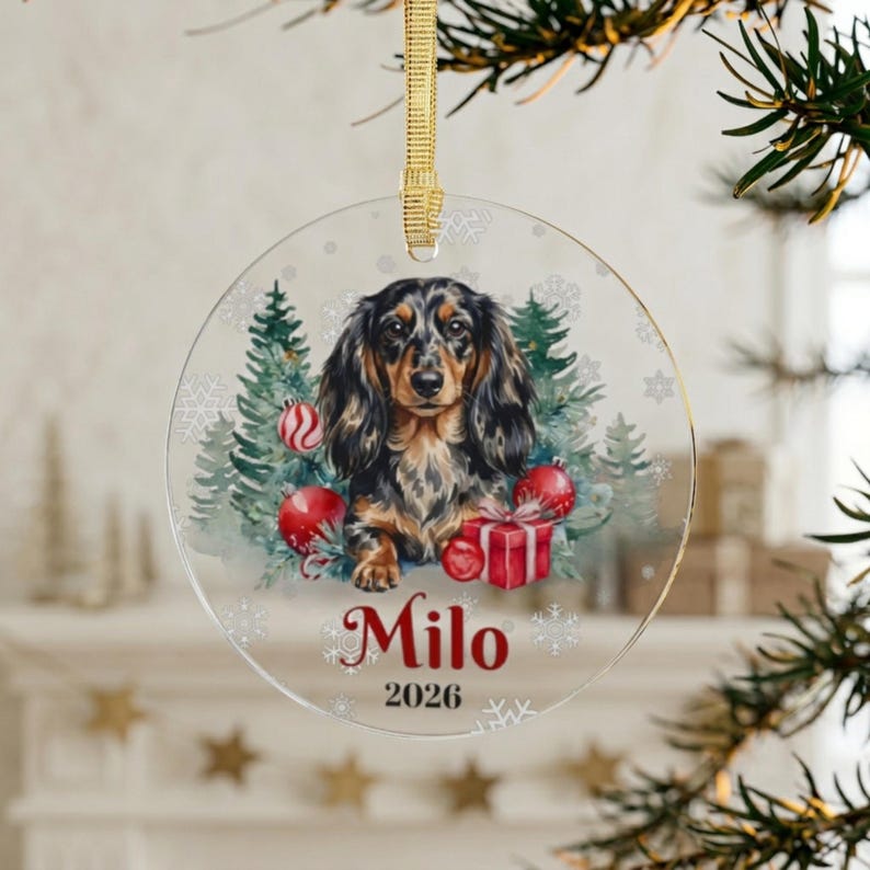 May include: A circular clear acrylic Christmas ornament featuring a long-haired dachshund illustration surrounded by green pine trees, red ornaments, and a gift box. The name Milo and the year 2026 are printed in red below the dog, with a gold ribbon.