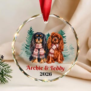 May include: A round, clear, faceted glass ornament hanging from a red ribbon. It features an illustration of two spaniel dogs wearing patterned scarves in front of green pine trees. The text reads Archie & Teddy and the year 2026.