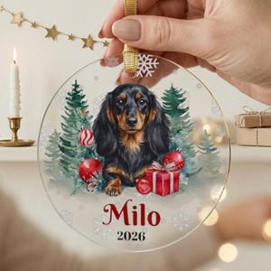 May include: A round, clear acrylic Christmas ornament featuring a long-haired black and tan dachshund illustration, green pine trees, red ornaments, and a gift box. The text "Milo" and "2026" is printed in red below the dog, with white snowflakes.