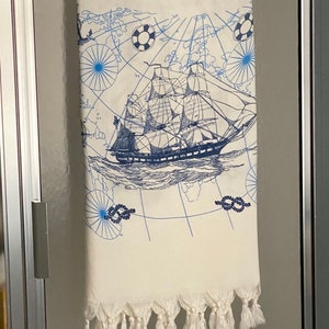 May include: Cream-colored nautical-themed towel with a detailed blue illustration of a sailing ship, nautical map, and decorative elements. The towel has a fringe along the bottom edge. The design evokes a vintage aesthetic.