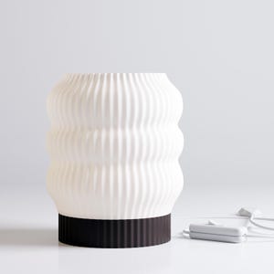 Könnte beinhalten: Eine weiße Tischlampe aus Keramik mit einem wellenförmigen, strukturierten Design und einem schwarzen Sockel. Die Lampe ist eingesteckt und betriebsbereit.
