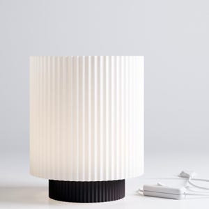 Puede incluir: Una lámpara de mesa blanca con una pantalla de tela plisada y una base negra. La lámpara está enchufada y el cable es visible.