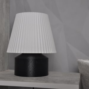 Könnte beinhalten: Ein weißer, gefalteter Lampenschirm sitzt auf einem schwarzen Lampenfuß. Die Lampe steht auf einer hellgrauen Oberfläche.