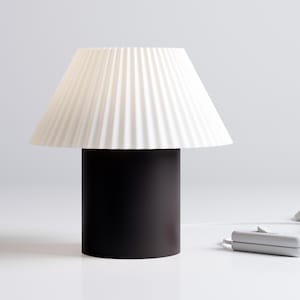 Może przedstawiać: Biały, plisowany klosz lampy na czarnej, cylindrycznej podstawie. Lampa stoi na białym tle.