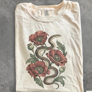 Op de afbeelding: Een ivoorkleurig t-shirt met een grafische print van een kronkelende slang verweven met grote rode klaprozen en groene bladeren. Het t-shirt heeft een ronde hals en een Comfort Colors merklabel aan de binnenzijde van de kraag.