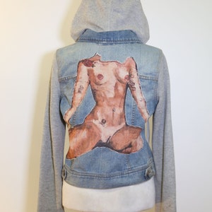 May include: A blue denim jacket with a gray hooded sweatshirt attached to the sleeves. The back of the jacket features a nude figure with tattoos.