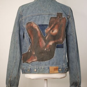 Peut inclure: Une veste en jean bleue avec une image peinte d'une figure nue au dos. La figure est brune et a une pose détendue.
