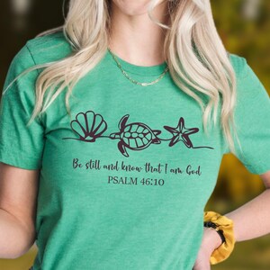May include: A green t-shirt featuring a seashell, turtle, and starfish design with the text "Be still and know that I am God" and "Psalm 46:10" in black. The shirt is made of a soft material.