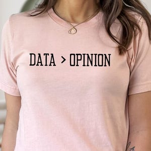 May include: A light pink t-shirt with the text "DATA > OPINION" in black block letters. The shirt is short-sleeved and made of a soft material. The model is wearing blue jeans.
