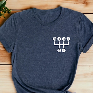 May include: A dark blue t-shirt with a white graphic of a 5-speed manual transmission gear shift pattern. The graphic includes the letters R, 1, 2, 3, 4, and 5.