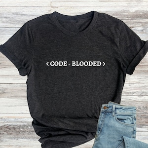 Könnte beinhalten: Ein dunkelgraues T-Shirt mit dem Text "<CODE-BLOODED>" in weiß auf der Vorderseite. Das T-Shirt ist gefaltet und auf einer Holzoberfläche mit einer blauen Jeans abgelegt.