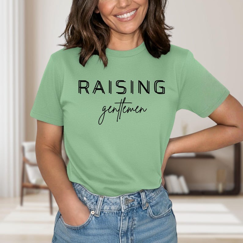 May include: A light green t-shirt with the text "RAISING gentlemen" in black. The word "RAISING" is in a bold, sans-serif font, while "gentlemen" is in a cursive script. The shirt is made of a soft material.