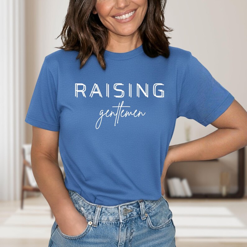 May include: A blue t-shirt with the white text "RAISING gentlemen" in a block and script font. The shirt is short-sleeved and made of a soft material. The person wearing the shirt is smiling.