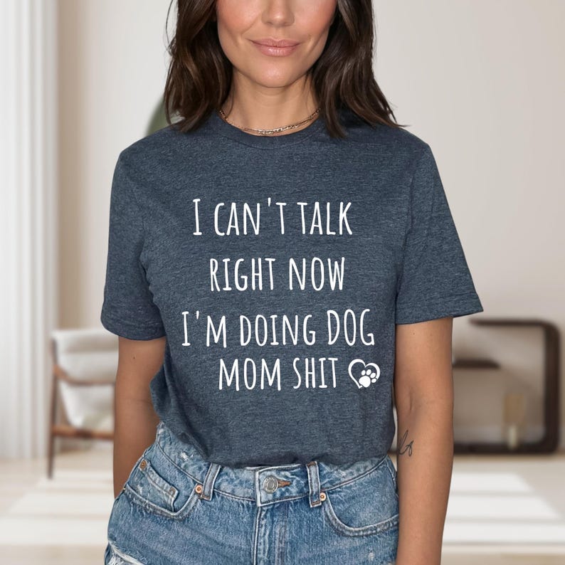 May include: A dark gray t-shirt with the text "I CAN'T TALK RIGHT NOW I'M DOING DOG MOM SHIT" in white. A paw print graphic is below the text. The shirt is paired with blue denim jeans.