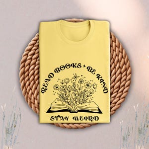 May include: A yellow t-shirt with the text "Read Books + Be Kind" arched above an open book illustration filled with flowers. Below the book is the text "Stay Weird". The shirt is displayed on a woven, circular placemat.
