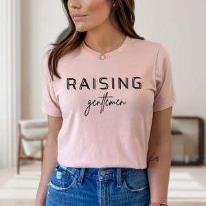 May include: A light pink t-shirt with the words "RAISING gentlemen" printed in black. The word "RAISING" is in a bold, sans-serif font, while "gentlemen" is in a cursive script. The shirt is made of a soft material.