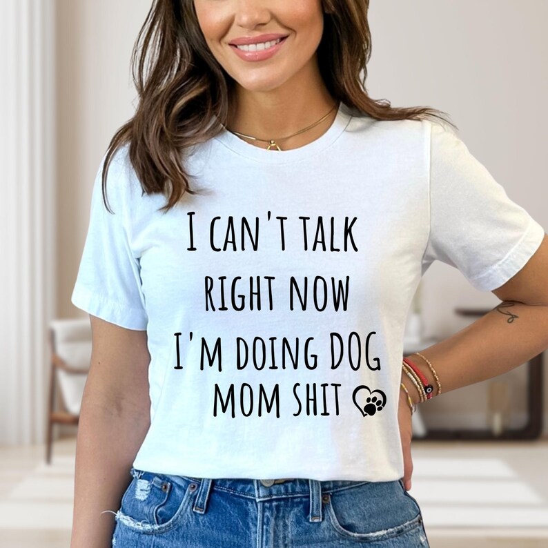 May include: White t-shirt with the text "I CAN'T TALK RIGHT NOW I'M DOING DOG MOM SHIT" in black. The design includes a heart and paw print graphic. The shirt is worn by a person in blue jeans.