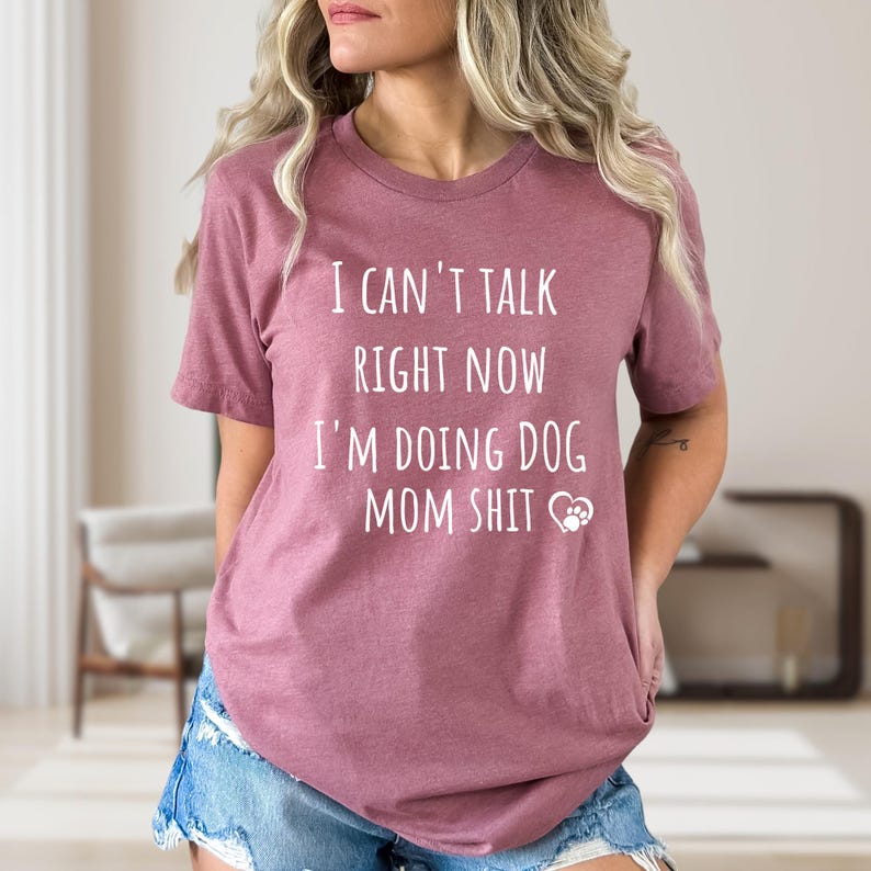 May include: A mauve-colored t-shirt with the text "I CAN'T TALK RIGHT NOW I'M DOING DOG MOM SHIT" in white block letters. A white paw print graphic is below the text. The shirt has short sleeves.