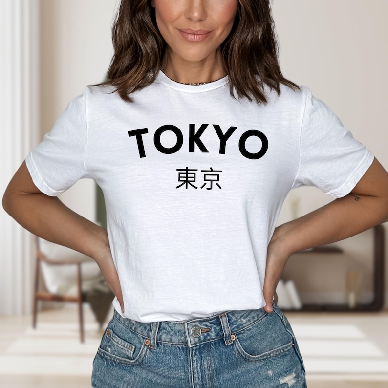 May include: White t-shirt with the word "TOKYO" in bold black letters above Japanese characters. The shirt is a crew neck style and is being worn with blue jeans.