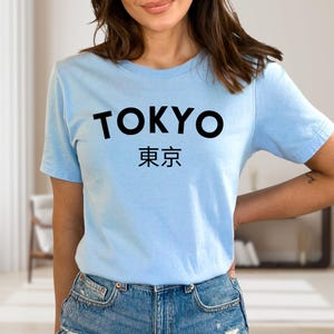 May include: Light blue t-shirt with the word "TOKYO" in bold black letters above Japanese characters. The shirt is made of a soft material and is suitable for casual wear.
