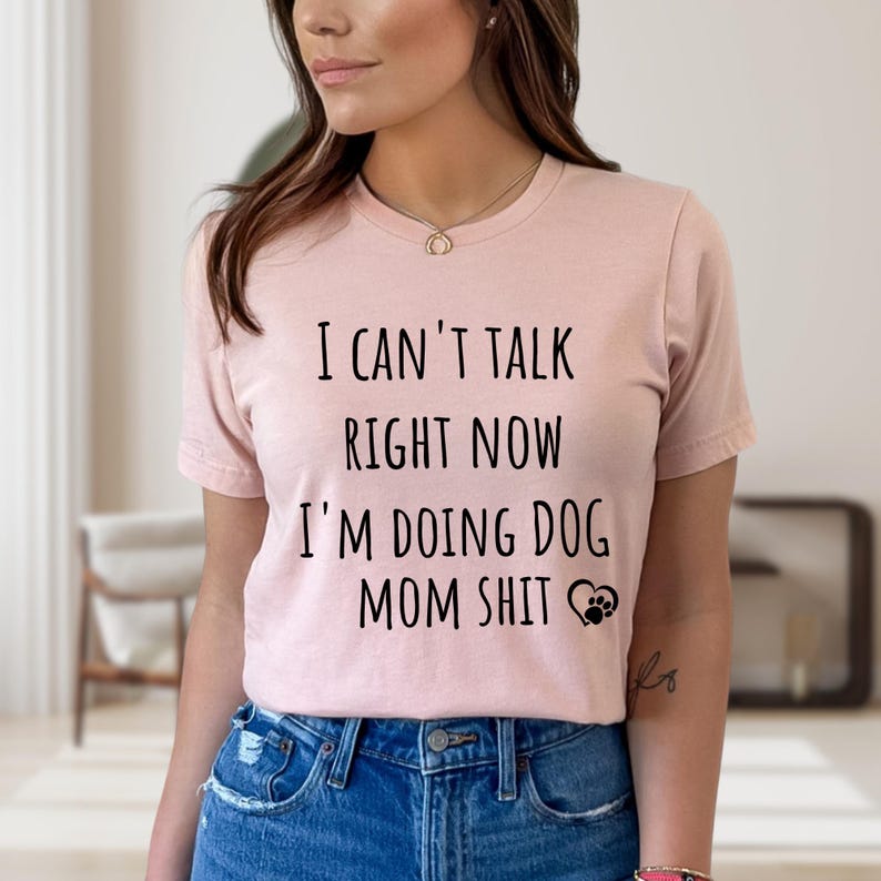May include: A light pink t-shirt with the text "I CAN'T TALK RIGHT NOW I'M DOING DOG MOM SHIT" in black. A paw print and heart graphic is below the text. The shirt is paired with blue jeans.