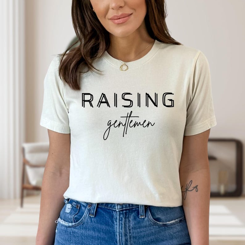 May include: Cream-colored t-shirt with the words "RAISING gentlemen" printed in black. The top text is in a bold, sans-serif font, while the word "gentlemen" is in a cursive script. The shirt is paired with blue jeans.
