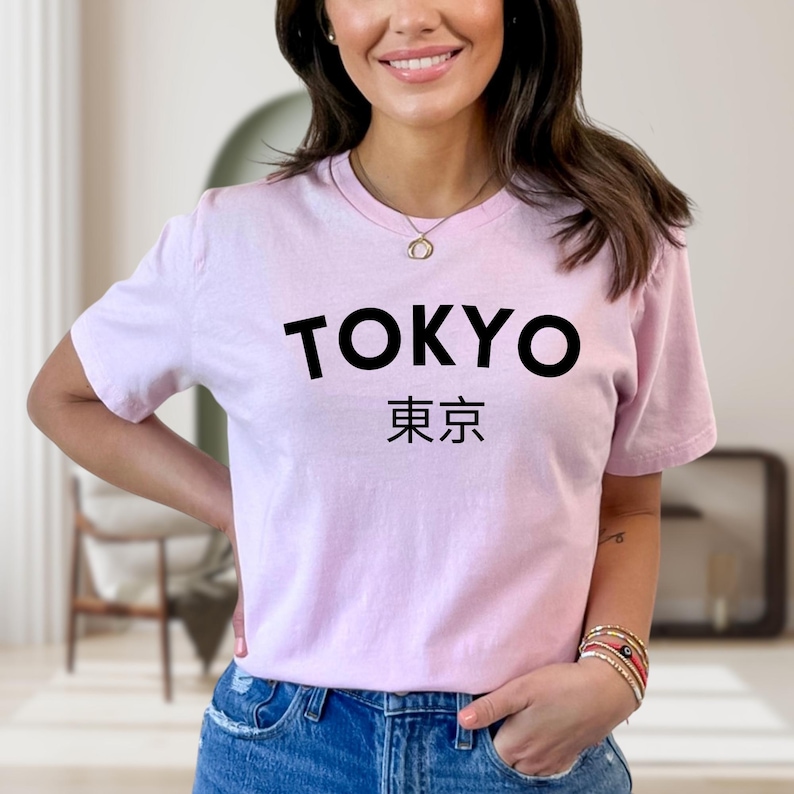 May include: A light pink t-shirt with the word "TOKYO" in bold black letters above Japanese characters. The shirt is a crew neck style and is made of a soft material.
