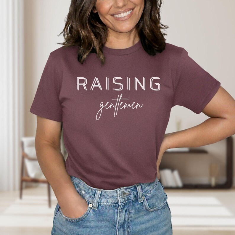 May include: A maroon t-shirt with the words "RAISING gentlemen" printed in white. The top is a crew neck style and is paired with blue jeans. The text is in a sans-serif font above a cursive font.