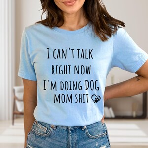 May include: Light blue t-shirt with the text "I CAN'T TALK RIGHT NOW I'M DOING DOG MOM SHIT" in black. A paw print graphic is next to the text. The shirt is made of a soft material.