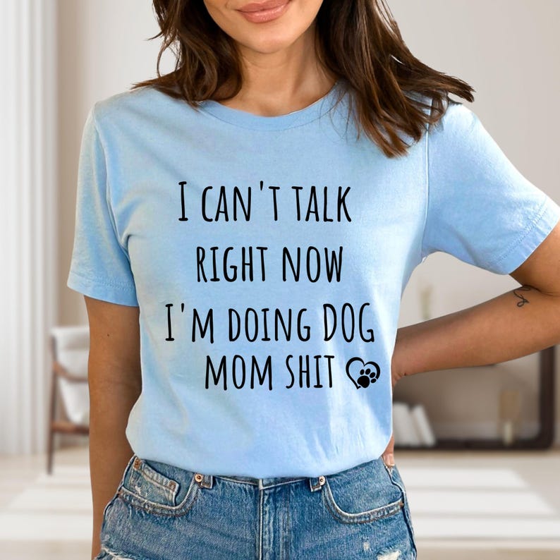 May include: Light blue t-shirt with the text "I CAN'T TALK RIGHT NOW I'M DOING DOG MOM SHIT" in black. A paw print graphic is next to the text. The shirt is made of a soft material.