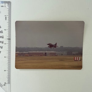 May include: A black and white military jet plane taking off from a runway. The plane is in the air and the runway is in the foreground. The sky is cloudy and the background is a field of grass.