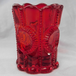 May include: A red glass toothpick holder with a scalloped rim and a repeating pattern of circles. The holder is made of clear glass with a red tint.