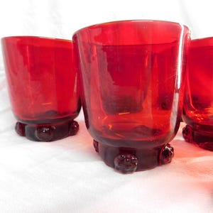 May include: Four red glass tumblers with a textured, rounded base. The glasses are clear and have a slightly frosted appearance.