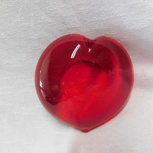 May include: A heart-shaped, solid red glass paperweight. The transparent glass has a glossy finish, reflecting light. The heart is resting on a white, textured surface. A decorative item.
