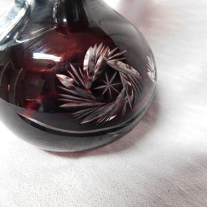 May include: A dark red glass decorative vase with a round body and a small handle. The vase features intricate cut glass designs, including a starburst pattern and leaf-like shapes. The glass is highly reflective, creating a play of light and shadow.