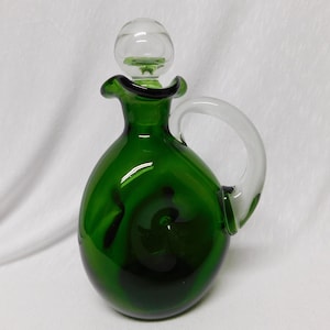 May include: A green glass decanter with a clear glass stopper and a handle. The decanter has a rounded body and a textured surface.