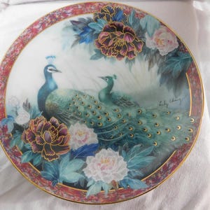 May include: A decorative ceramic plate with a colorful painting of two peacocks with their tails spread out. The plate features a floral design with pink, white, and purple flowers. The plate is trimmed in gold and has the artist's signature in the bottom right corner.